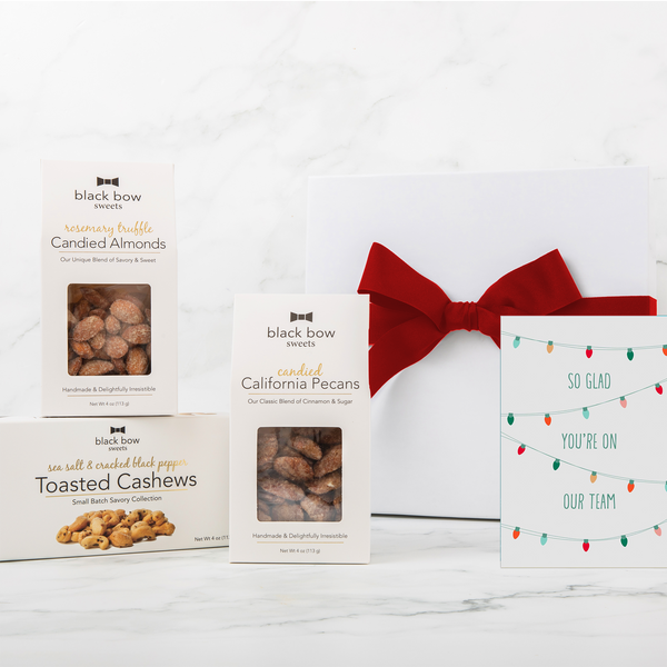 A Black Bow Sweets gift box with a large red bow next to packages of candied California cinnamon and sugar coated pecans, candied rosemary truffle almonds, and toasted sea salt and cracked black pepper cashews next to a gift enclosure card picturing Christmas lights that says "So glad you're on our team".
