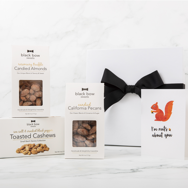 A Black Bow Sweets gift box with a large black bow next to packages of candied California cinnamon and sugar coated pecans, candied rosemary truffle coated almonds, and sea salt and cracked black pepper toasted cashews next to a gift enclosure card picturing a squirrel holding a nut that says "I'm nuts about you".