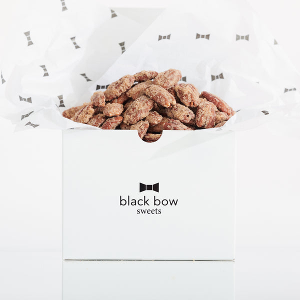 A white Black Bow Sweets gift box filled with candied California cinnamon and sugar pecans loosely wrapped in white tissue paper with small black bows.