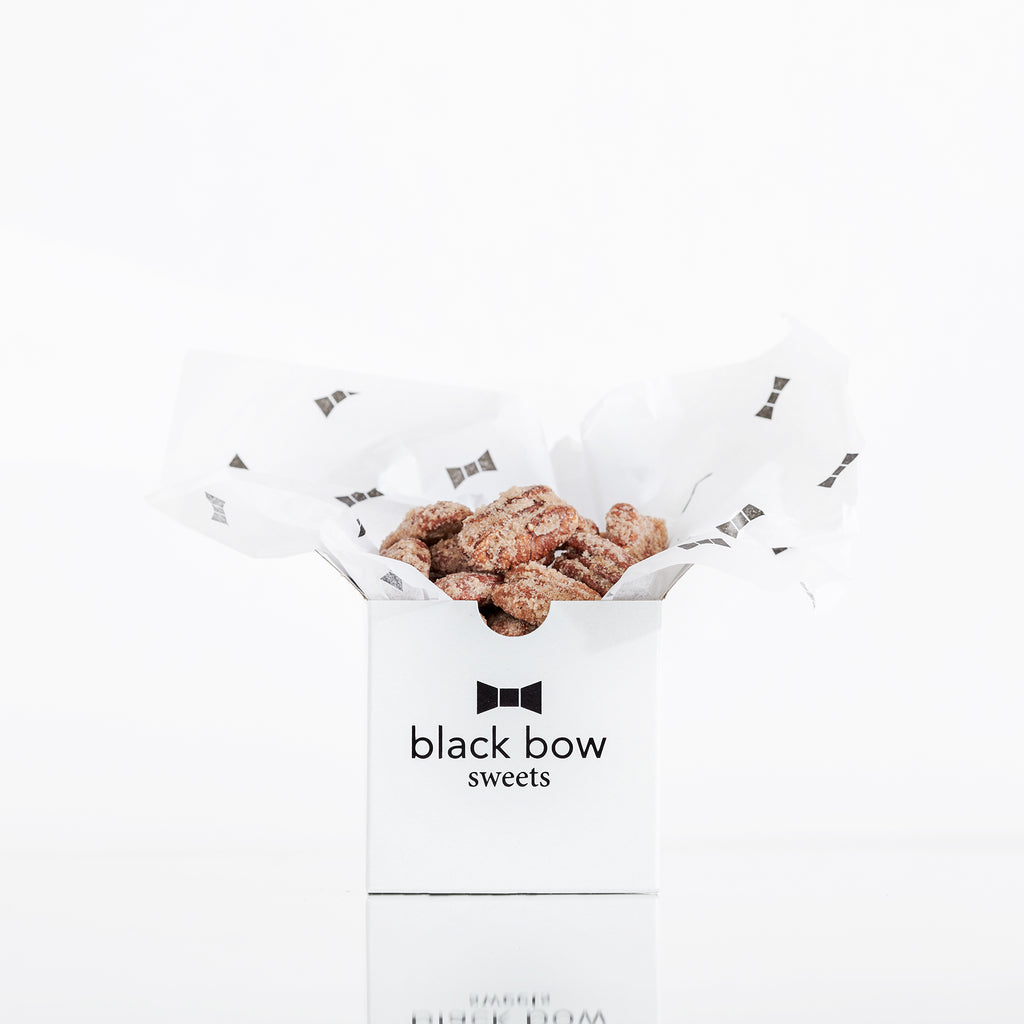 A Black Bow Sweets mini gift box filled with candied California cinnamon and sugar coated pecans loosely wrapped in white tissue paper with small black bows.