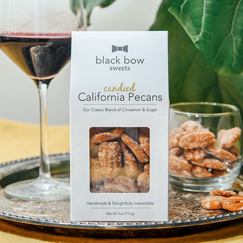 A package of Black Bow Sweets candied California cinnamon and sugar coated pecans on a silver platter next to a glass filled with the pecans and a glass of red wine.