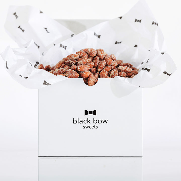 A white Black Bow Sweets gift box filled with candied cinnamon and sugar almonds loosely wrapped in white tissue paper with small black bows.
