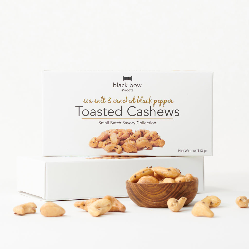 A box of Black Bow Sweets sea salt and cracked pepper toasted cashews next to a wooden bowl filled with the cashews.