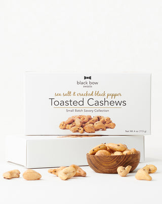 A box of Black Bow Sweets sea salt and cracked black pepper toasted cashews next to a wooden bowl filled with the cashews.