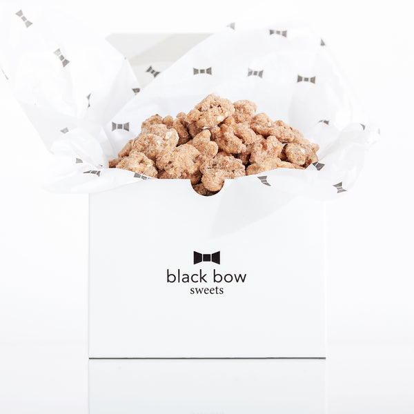 A white Black Bow Sweets gift box filled with candied California cinnamon and sugar coated walnuts loosely wrapped in white tissue paper with small black bows.