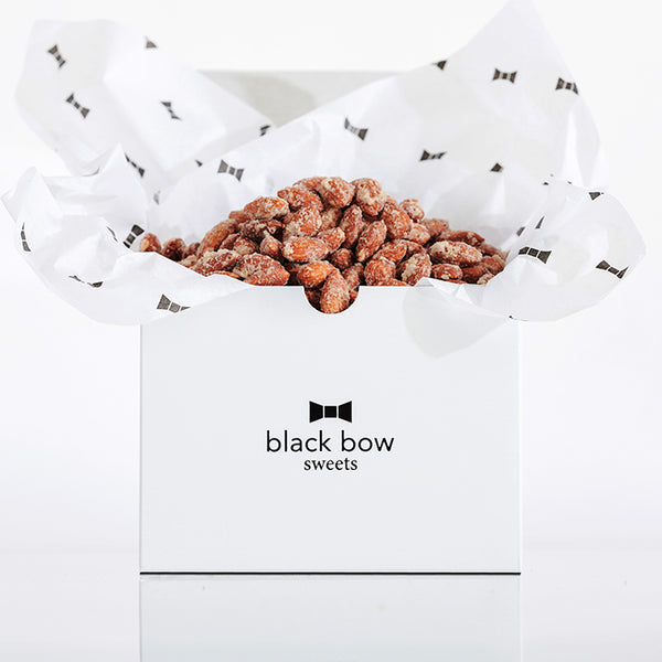 A white Black Bow Sweets gift box filled with candied cinnamon and sugar almonds loosely wrapped in white tissue paper with small black bows.